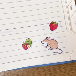 May include: A notebook with a blue cover and white pages. There are three stickers on the pages: a red strawberry, a green strawberry with a white flower, and a brown mouse.