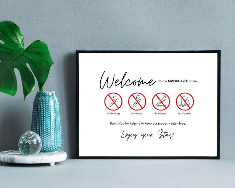 Op de afbeelding: Een ingelijst bord met de woorden "Welcome" en "Enjoy your Stay!" in zwart schrift. Het bord heeft vier rode cirkeliconen met een streep erdoor, die "No Smoking", "No Vaping", "No Incense" en "No Candles" aangeven. De tekst "SMOKE-FREE home" is ook opgenomen.