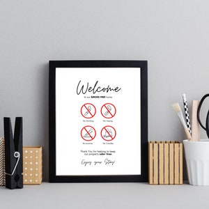 Op de afbeelding: Een ingelijst bord met een zwarte rand toont de woorden "Welcome" en "to our SMOKE FREE home." Hieronder staan vier rode en zwarte verbodssymbolen voor roken, vapen, wierook en kaarsen. Het bord bevat ook de tekst "Thank You for helping to keep our property odor-free. Enjoy your Stay!"