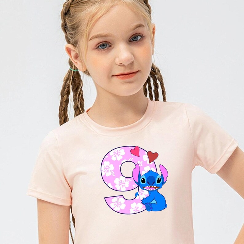 May include: A pink t-shirt with a blue Stitch character holding a pink number nine with white hibiscus flowers. The number nine is surrounded by two red hearts.