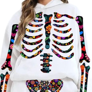 May include: White hoodie featuring a colorful floral skeleton design. The skeleton's bones are decorated with vibrant flowers, hearts, and patterns in various colors. The design covers the front of the hoodie, creating a unique and eye-catching look.