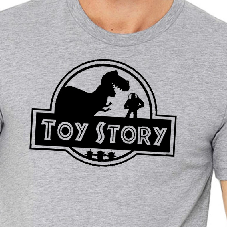 Może przedstawiać: Szary T-shirt z czarnym nadrukiem logo Jurassic Park, ale z napisem "Toy Story" zamiast "Jurassic Park". Grafika przedstawia sylwetkę dinozaura i postać kosmicznego strażnika.