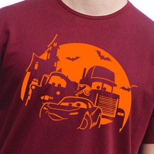 May include: Burgundy t-shirt featuring an orange Halloween-themed graphic. The design includes cartoon cars, a haunted house, bats, and a full moon. The graphic is centered on the shirt, creating a festive and eye-catching design.