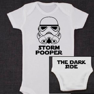 May include: White baby onesie with a black and white image of a Stormtrooper helmet and the text "Storm Pooper". The onesie has a black and white text "The Dark Side" on the bottom.