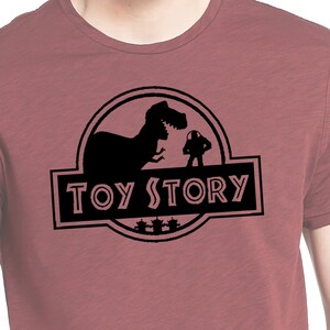 Może przedstawiać: Koszulka w kolorze bordowym z czarną sylwetką dinozaura i postacią z filmu Toy Story. Tekst "TOY STORY" jest nadrukowany na czarnym banerze pod sylwetką.