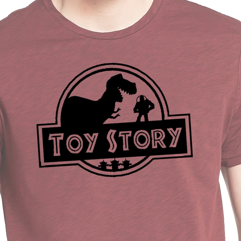 Może przedstawiać: Koszulka w kolorze bordowym z czarną sylwetką dinozaura i postacią z filmu Toy Story. Tekst "TOY STORY" jest nadrukowany na czarnym banerze pod sylwetką.