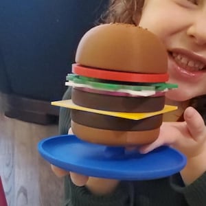 May include: A large, toy hamburger with a sesame seed bun, red tomato, green lettuce, purple onion, and yellow cheese. The burger sits on a blue plate. The burger is held by a person.