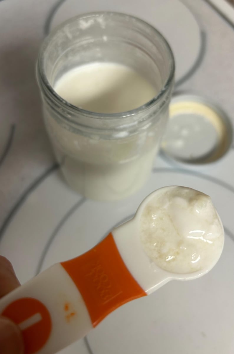May include: A white measuring spoon filled with a thick, creamy white substance. The spoon is orange and white and has a number "1" on the side.