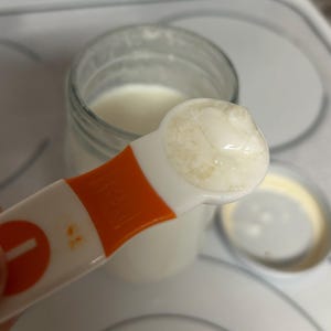May include: A white measuring spoon filled with a thick, white yogurt-like substance. The spoon is orange and white and has a number "1" on the handle.