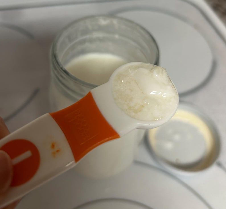 May include: A white measuring spoon filled with a thick, white yogurt-like substance. The spoon is orange and white and has a number "1" on the handle.