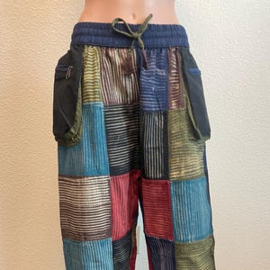 Może przedstawiać: Spodnie w stylu patchwork z niebieskim ściągaczem w pasie i dwiema czarnymi kieszeniami bocznymi. Spodnie posiadają różnorodne kolorowe prostokątne łaty w odcieniach niebieskiego, zielonego, czerwonego i brązowego. Tkanina ma wzór w paski.
