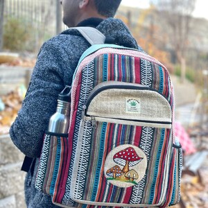 May include: A multi-coloured striped hemp backpack with a mushroom patch and a small hemp patch that says "Hemp Incense" on it. The backpack has a side pocket and a zipped front pocket.