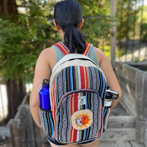May include: A multi-colored striped hemp backpack with a sun and moon design. The backpack has a large main compartment and two side pockets. The backpack is made of hemp and is a sustainable and eco-friendly option.