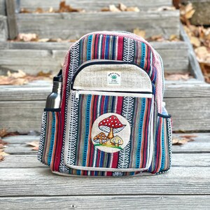 May include: A multi-coloured striped hemp backpack with a large embroidered patch of three mushrooms. The patch is in a circle with a white background and the mushrooms are red, white, and brown. The backpack has a zipped pocket on the front and a small hemp patch with the text "Himalayan Hemp The Tree Free Alternative" on the front.