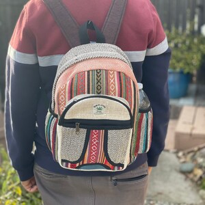 Könnte beinhalten: Ein weißer Hanfrucksack mit bunten Streifen und einer kleinen Tasche auf der Vorderseite. Der Rucksack hat einen schwarzen Reißverschluss und ein kleines grünes Etikett mit der Aufschrift "Himalaya Hemp".