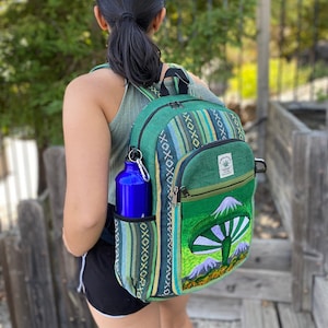 May include: A green and blue backpack with a mushroom design and the text "Herbal Hemp" on the front. The backpack has a side pocket with a blue water bottle.