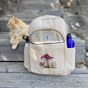 May include: A beige hemp backpack with a zippered pocket and a mushroom embroidery design. The backpack has the text "Himalayan Hemp" and "Made in Nepal" on a brown tag.