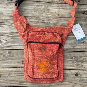 May include: A red canvas fanny pack with a tree of life design and a zippered pocket. The bag has a long adjustable strap with a buckle.