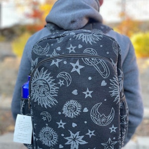 Może przedstawiać: Czarny plecak z płótna z nadrukiem kosmicznym przedstawiającym słońca, księżyce, gwiazdy i serca. Nadruk jest w jasnoszarym kolorze.
