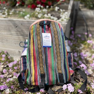 May include: A colorful backpack with a striped pattern in red, yellow, green, and blue. The backpack has black straps and a side pocket. A water bottle is visible on the side. The tag reads "Hand Made in Nepal".