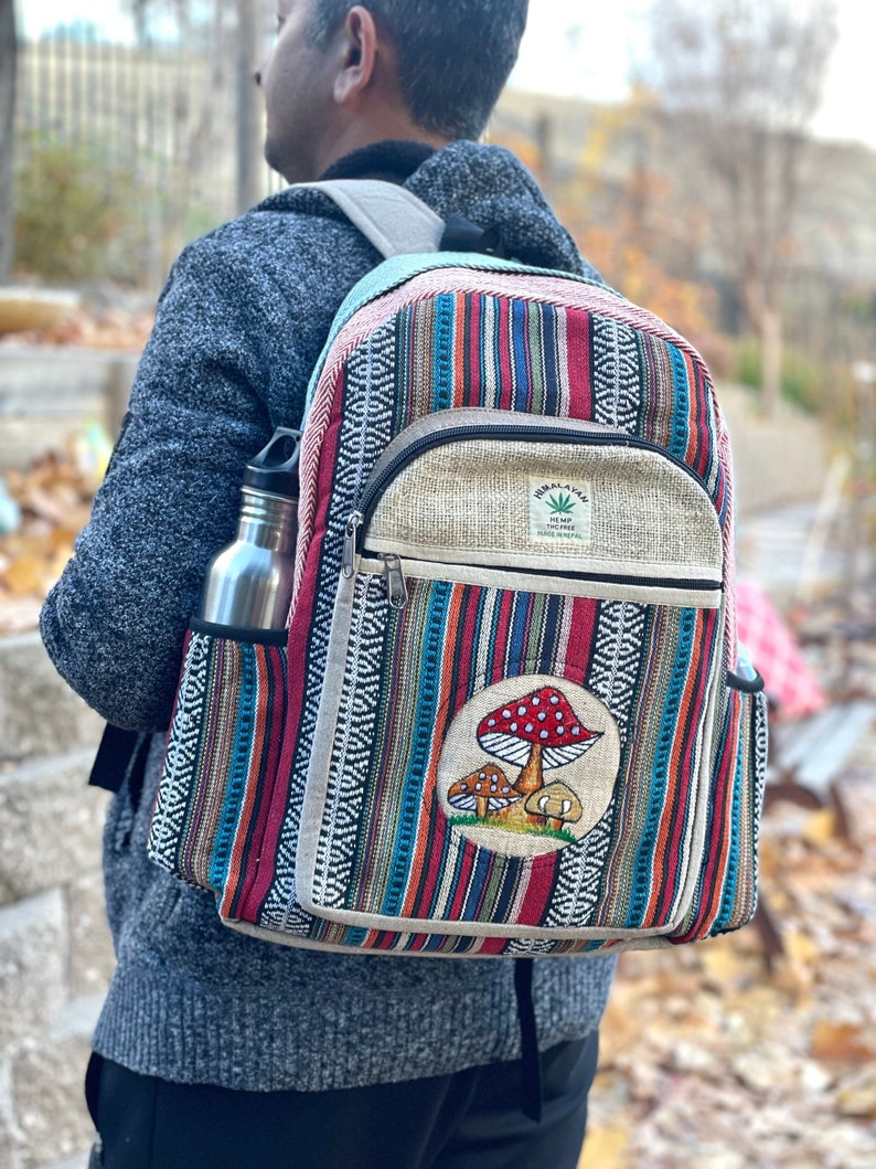 May include: A multi-coloured striped hemp backpack with a mushroom patch and a small hemp patch that says "Hemp Incense" on it. The backpack has a side pocket and a zipped front pocket.
