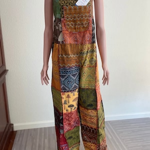 Può includere: Salopette in patchwork con un motivo multicolore che presenta forme geometriche, funghi e altri disegni. La salopette ha una grande tasca sul davanti e bretelle regolabili.