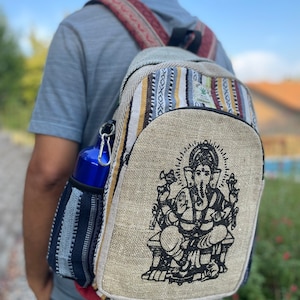 May include: A beige hemp backpack with a black and white image of the Hindu deity Ganesha on the front. The backpack has a multi-colored striped pattern on the top flap and a blue water bottle in the side pocket.