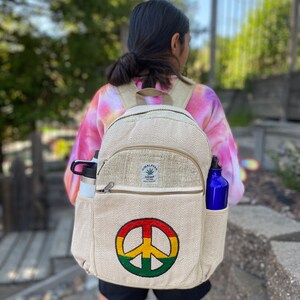 May include: A beige hemp backpack with a peace sign embroidered on the front. The backpack has a zippered pocket on the front and two side pockets. The backpack is made of natural hemp fabric and has a Himalayan Hemp label on the front.