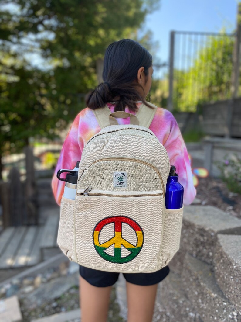 May include: A beige hemp backpack with a peace sign embroidered on the front. The backpack has a zippered pocket on the front and two side pockets. The backpack is made of natural hemp fabric and has a Himalayan Hemp label on the front.
