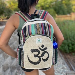 May include: A beige hemp backpack with a black Om symbol on the front. The backpack has a colorful striped pattern on the sides and a black strap. The backpack is made from Himalayan hemp.