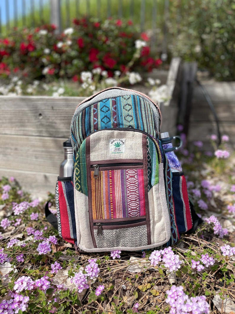 May include: A colorful backpack with a patchwork design, featuring various patterns and textures. The backpack has multiple pockets, including side pockets for water bottles. The overall color scheme includes blues, greens, reds, and browns, with a hemp leaf logo.