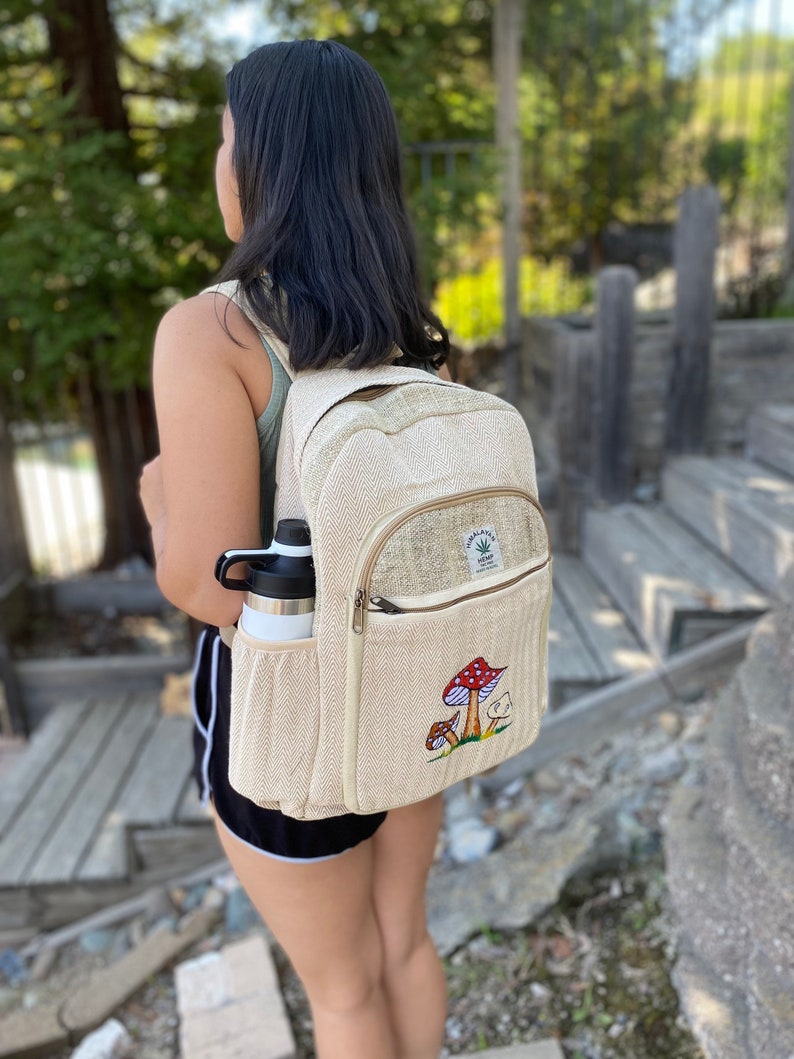 May include: A beige hemp backpack with a mushroom embroidery design. The backpack has a zippered pocket on the front and a side pocket for a water bottle. The text "Himalayan Hemp" is embroidered on the front of the backpack.