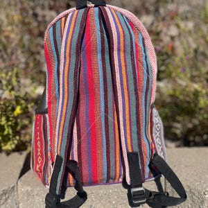 May include: A colorful backpack with vertical stripes in red, blue, orange, and green. The backpack has black straps and a black handle. The edges are trimmed with a pink and white patterned fabric. The backpack is sitting on a stone surface.