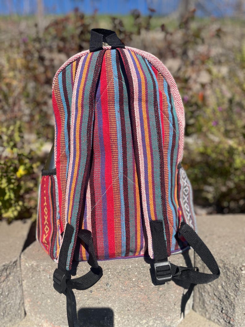 May include: A colorful backpack with vertical stripes in red, blue, orange, and green. The backpack has black straps and a black handle. The edges are trimmed with a pink and white patterned fabric. The backpack is sitting on a stone surface.