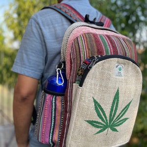 May include: A hemp backpack with a large green marijuana leaf design on the front. The backpack has a zippered pocket and a side pocket for a water bottle. The backpack is made of natural hemp fabric and has a colorful striped pattern on the side.