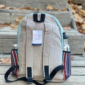May include: A beige canvas backpack with a blue and white striped trim. The backpack has a black adjustable strap and a small tag that says "LIKE INTERNATIONAL" with the style number "MI-257". The backpack has a side pocket with a colourful striped pattern.