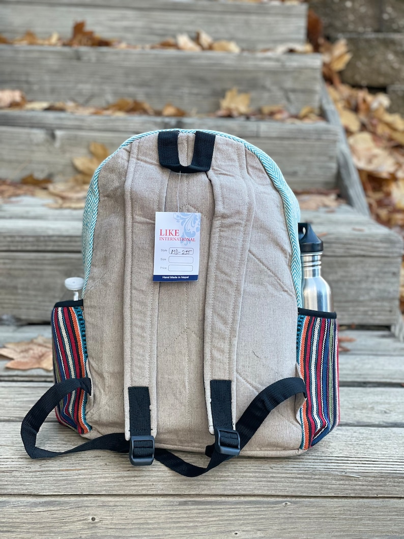 May include: A beige canvas backpack with a blue and white striped trim. The backpack has a black adjustable strap and a small tag that says "LIKE INTERNATIONAL" with the style number "MI-257". The backpack has a side pocket with a colourful striped pattern.