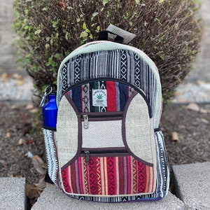 May include: A colorful backpack with a geometric pattern, featuring a blue water bottle in the side pocket. The backpack has multiple zippered compartments and a tag that reads "Himalaya Hemp". The bag is made of a woven fabric with a variety of colors.