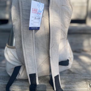 May include: A beige backpack with black adjustable straps and a tag that reads "LIKE INTERNATIONAL". The backpack has a quilted design on the back and shoulder straps. The tag also says "Hand Made in Nepal".
