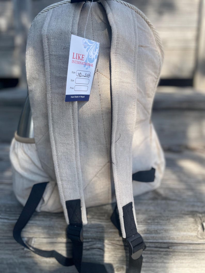 May include: A beige backpack with black adjustable straps and a tag that reads "LIKE INTERNATIONAL". The backpack has a quilted design on the back and shoulder straps. The tag also says "Hand Made in Nepal".