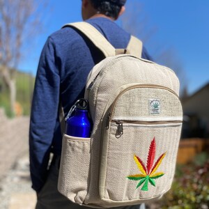 May include: A beige hemp backpack with a large green, yellow, and red marijuana leaf embroidered on the front. The backpack has a zippered pocket on the front and a side pocket that holds a blue water bottle. The backpack has a label that reads "Hemp Tailor" and "Sustainable Fashion".