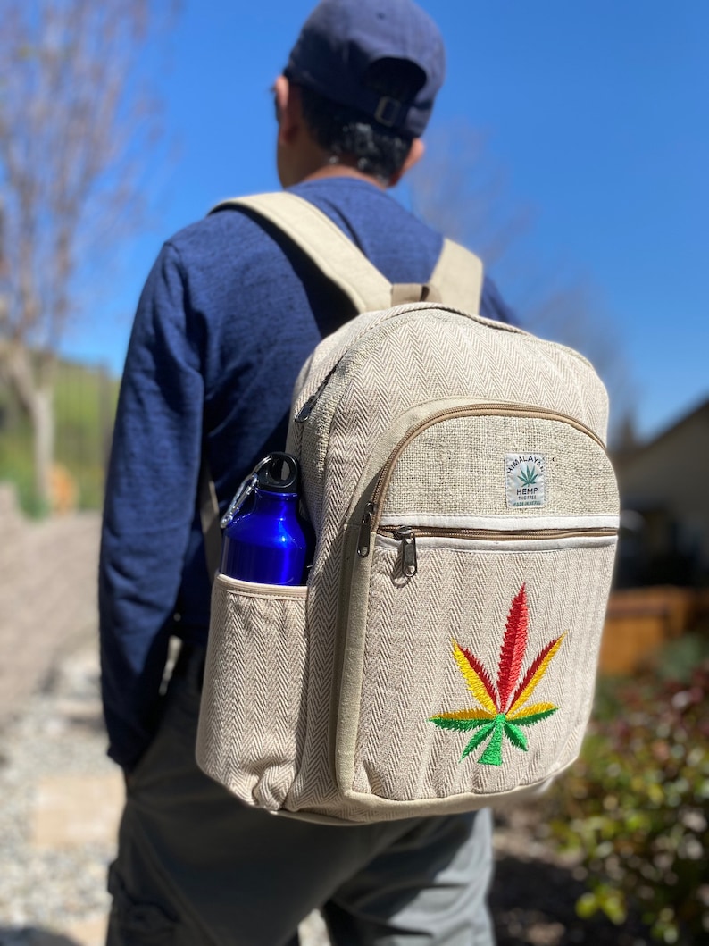 May include: A beige hemp backpack with a large green, yellow, and red marijuana leaf embroidered on the front. The backpack has a zippered pocket on the front and a side pocket that holds a blue water bottle. The backpack has a label that reads "Hemp Tailor" and "Sustainable Fashion".