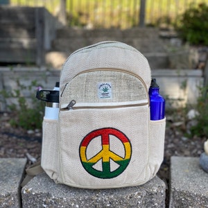 May include: A beige hemp backpack with a peace sign embroidered on the front. The backpack has a zippered pocket and two side pockets. The backpack is sitting on a stone surface.