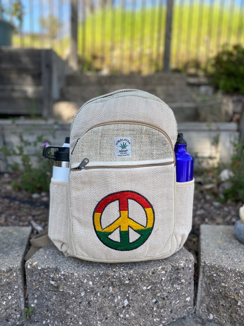 May include: A beige hemp backpack with a peace sign embroidered on the front. The backpack has a zippered pocket and two side pockets. The backpack is sitting on a stone surface.