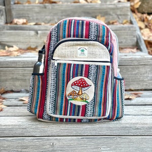 K&ouml;nnte beinhalten: Ein mehrfarbiger, gestreifter Hanfrucksack mit einem Pilz-Patch. Der Rucksack hat eine Rei&szlig;verschlusstasche vorne und eine Seitentasche f&uuml;r eine Wasserflasche. Der Text "HIMALAYAN HEAP THE TREE" befindet sich auf der vorderen Tasche.