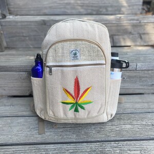 May include: A beige hemp backpack with a zippered pocket and a green, yellow, and red marijuana leaf embroidered on the front. The backpack has a brown leather patch with the text "Himalayan Hemp The Real Deal" on it.