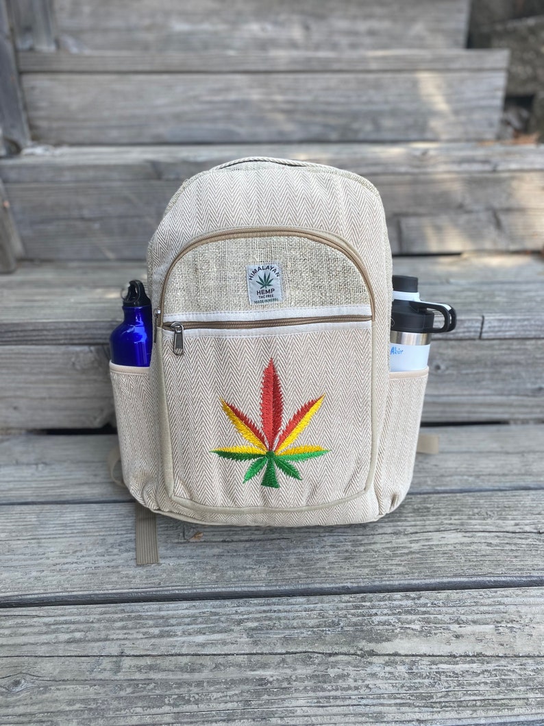 May include: A beige hemp backpack with a zippered pocket and a green, yellow, and red marijuana leaf embroidered on the front. The backpack has a brown leather patch with the text "Himalayan Hemp The Real Deal" on it.