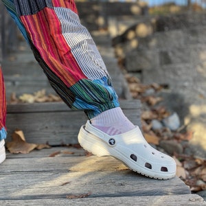 May include: White clogs with a textured sole and a small logo on the side. The clogs are worn by a person wearing colorful striped pants.