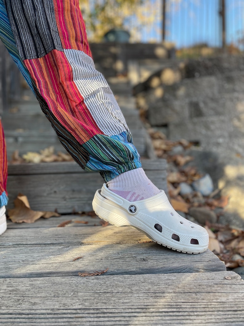 May include: White clogs with a textured sole and a small logo on the side. The clogs are worn by a person wearing colorful striped pants.