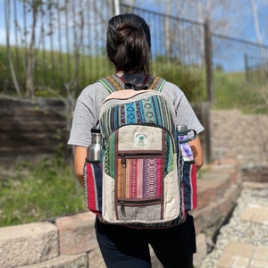May include: A colorful backpack with a hemp leaf logo, featuring a mix of woven patterns in blue, red, and brown. It has side pockets holding a silver water bottle and a purple-wrapped item. The backpack is worn by a person in a gray shirt and black pants.